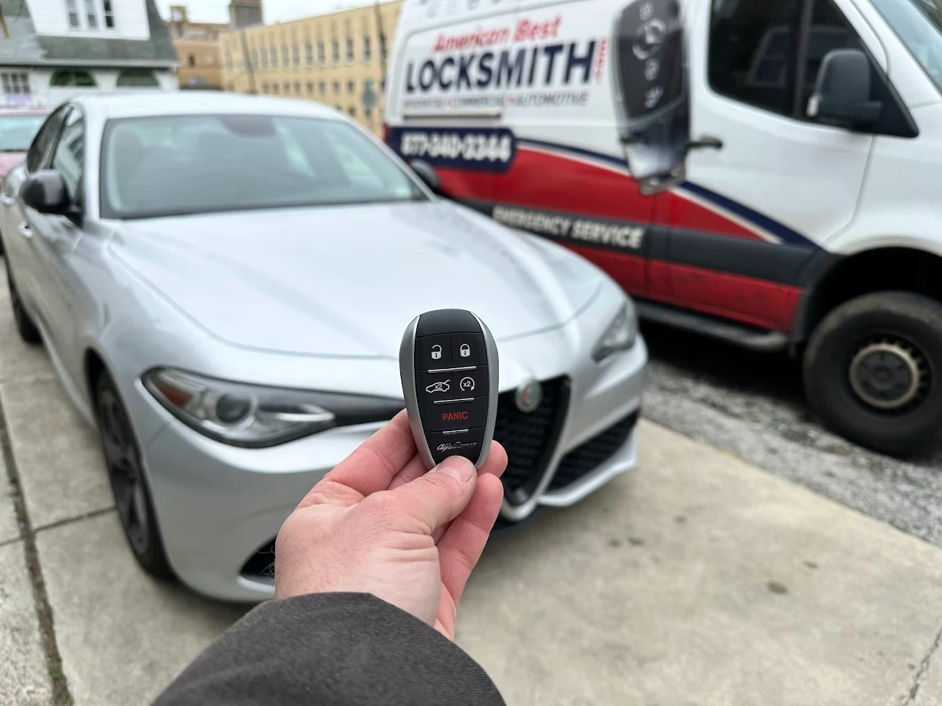 Freshly programmed Alfa Romeo smart key with the iconic Alfa shield emblem held in front of a silver Alfa Romeo Giulia with the American Best Locksmith service van visible behind