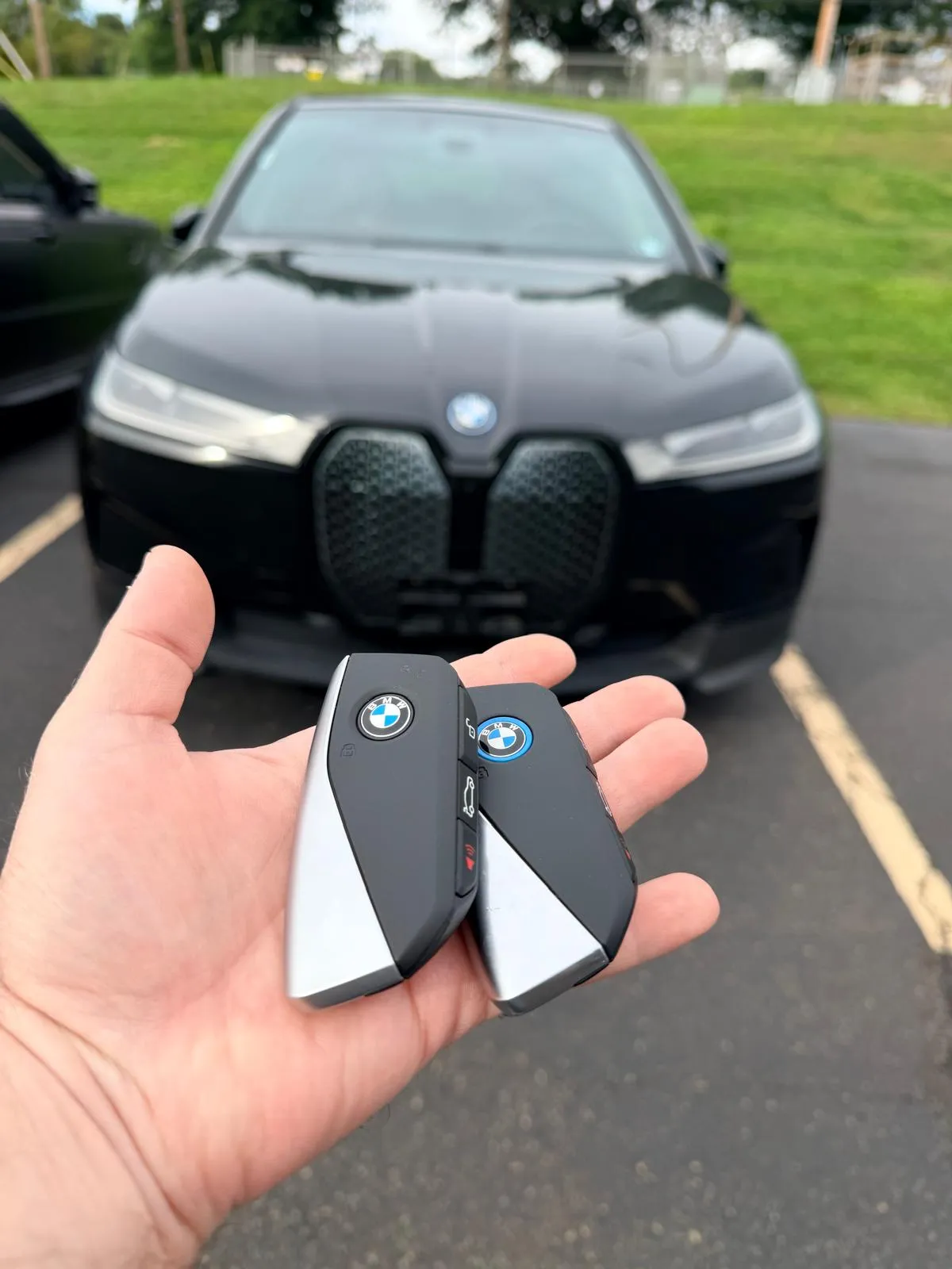 Two BMW smart keys held in front of a black 2023 BMW iX electric SUV in a parking lot after an on-site spare key programming
