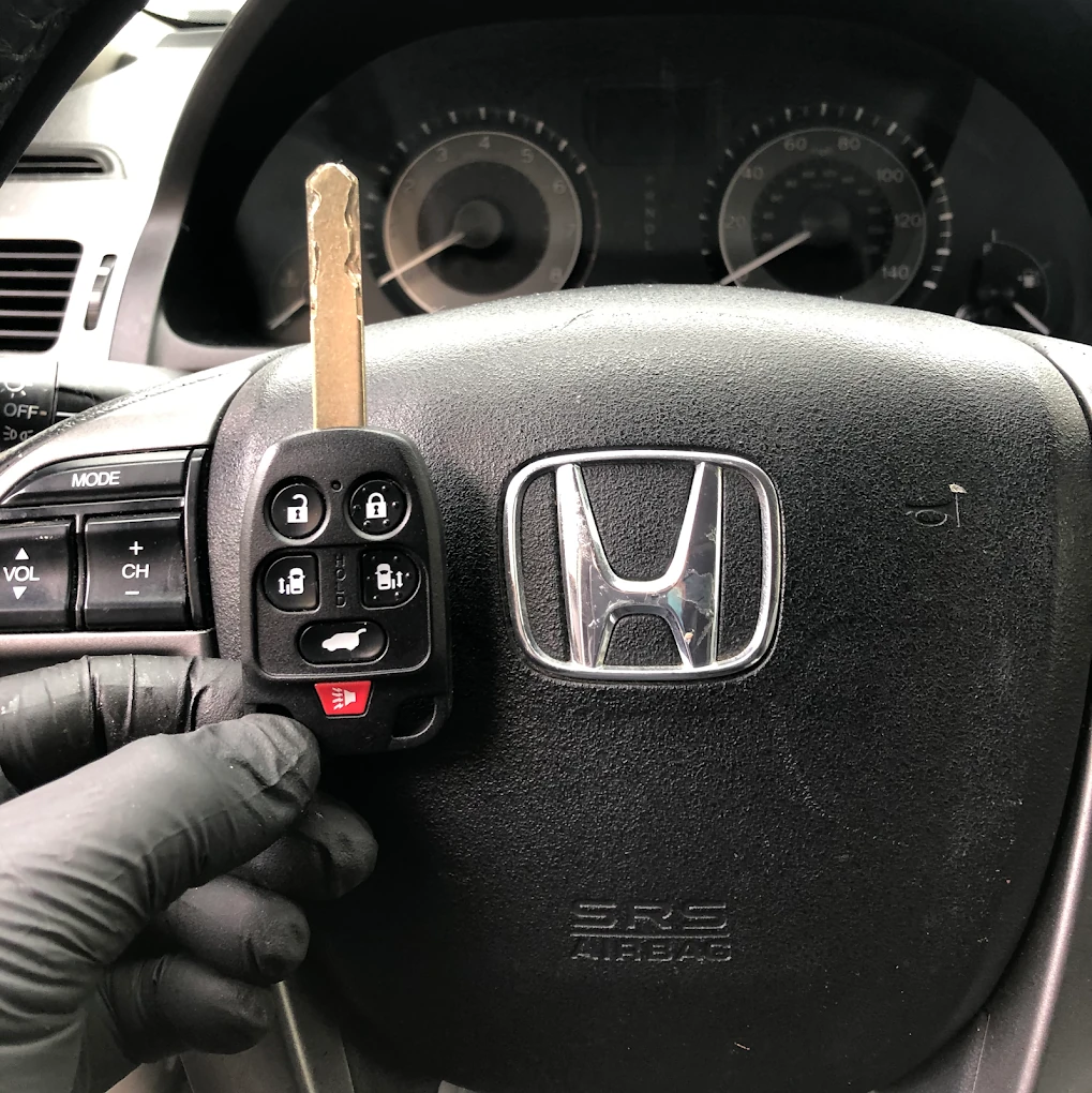 Freshly programmed Honda Odyssey 5-button remote with mechanical key blade held in gloved hand in front of the Honda steering wheel emblem and dashboard gauges
