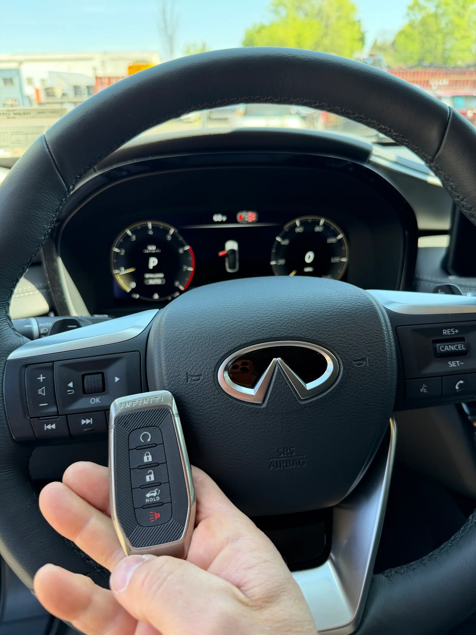 Freshly programmed 2025 Infiniti QX80 slim smart key held in front of the Infiniti steering wheel emblem with the new flagship's digital instrument cluster visible