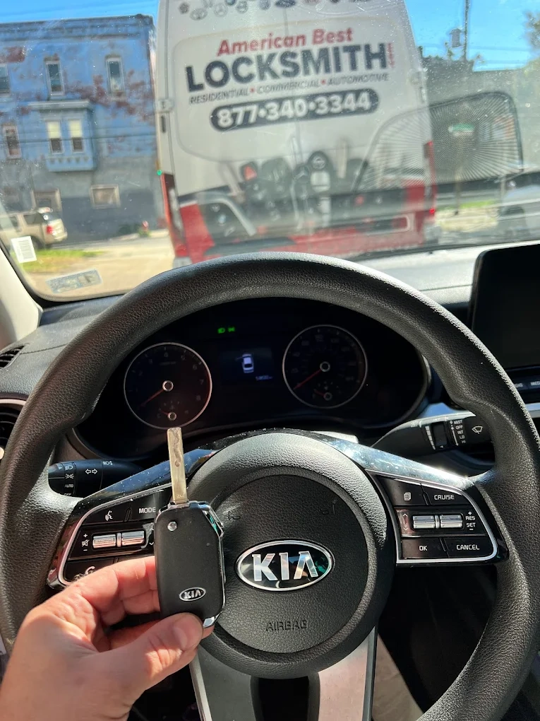 Freshly programmed Kia mechanical transponder key held in front of the Kia steering wheel airbag emblem with the American Best Locksmith service van visible through the windshield