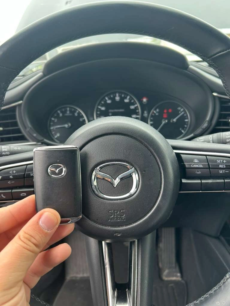 Freshly programmed 2025 Mazda CX-5 slim smart key held in front of the Mazda M-wing emblem on the steering wheel airbag with the modern analog instrument cluster visible behind