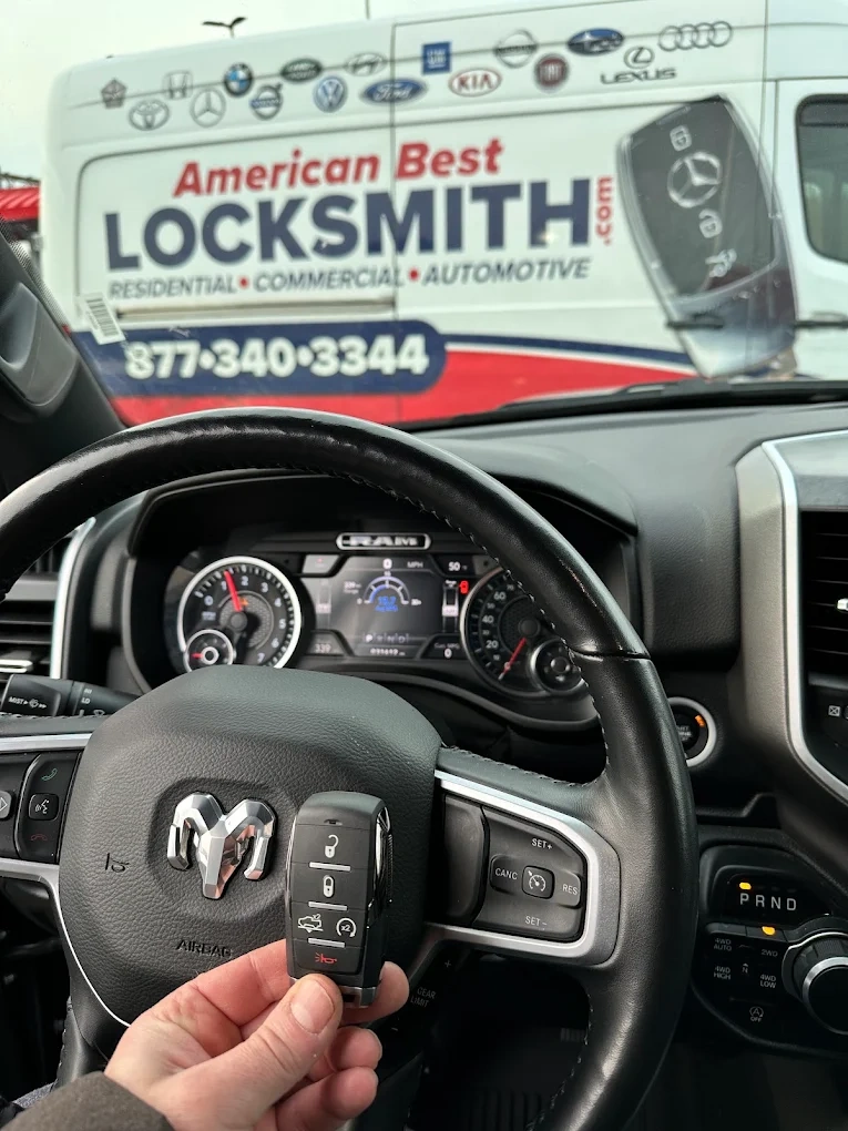 Freshly programmed RAM 1500 smart key held in front of the RAM head emblem on the steering wheel with the American Best Locksmith service van visible through the windshield showing all major brand emblems we service