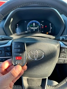 Freshly programmed 2024 Toyota Highlander Hybrid smart key held in front of the current-generation Toyota steering wheel emblem with the hybrid digital instrument cluster visible behind