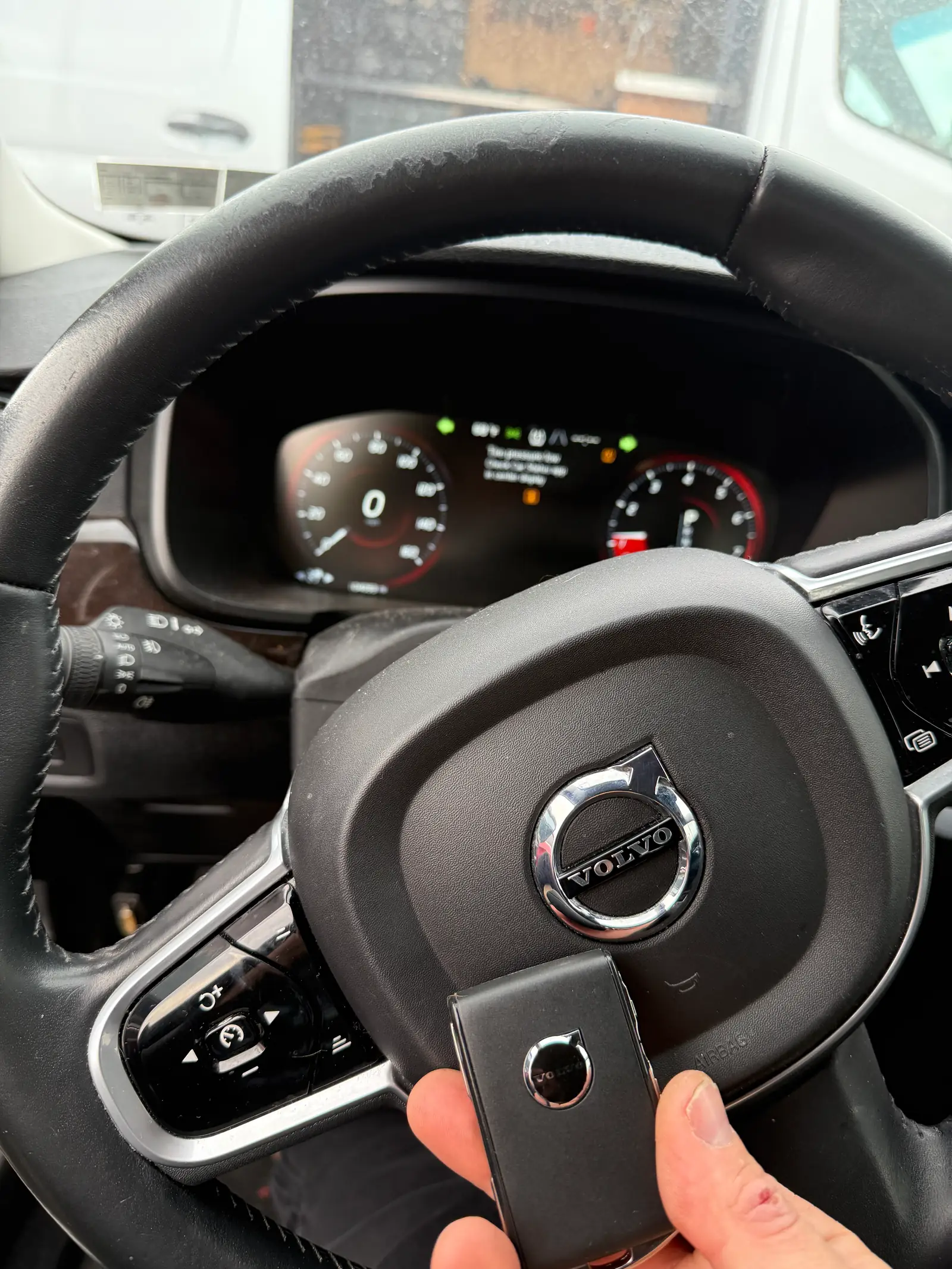 Freshly programmed 2025 Volvo slim smart key held in front of the Volvo Iron Mark emblem on the steering wheel airbag with the digital instrument cluster visible